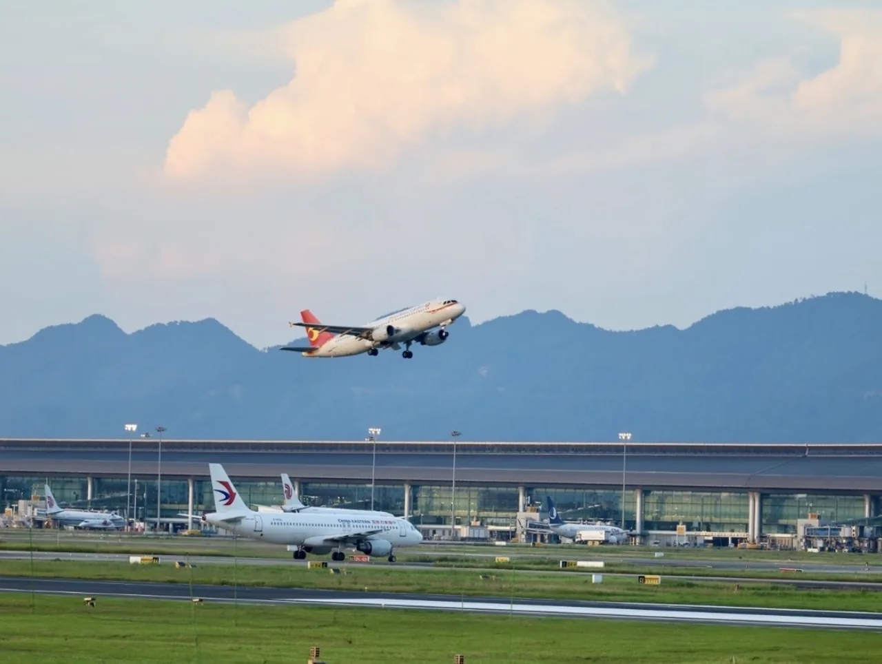 An aircraft taking off above an airport terminal with mountains in the background